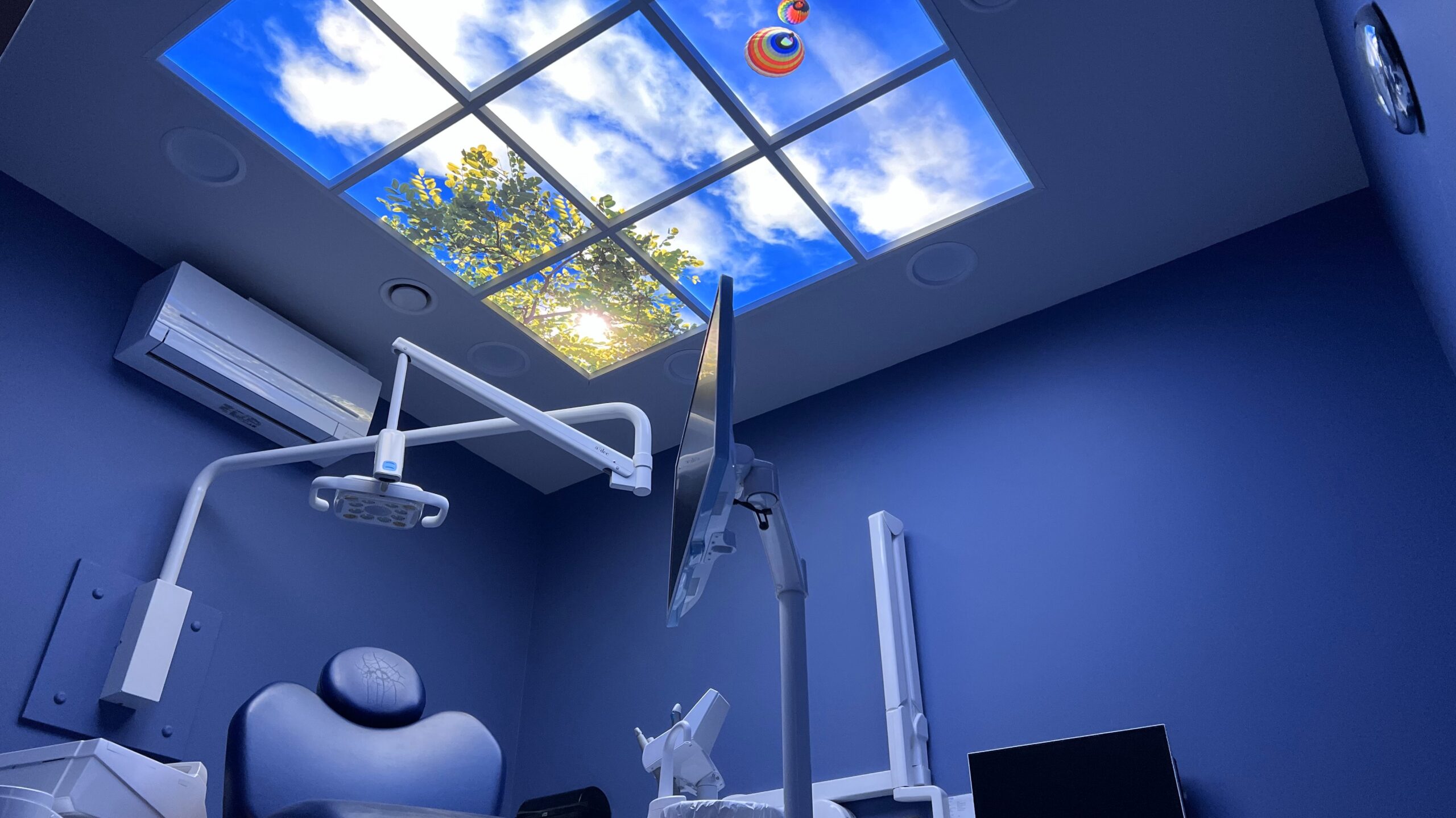 Tandartsbehandelkamer met een LED-fotoplafond dat een blauwe lucht met wolken en boomtakken toont